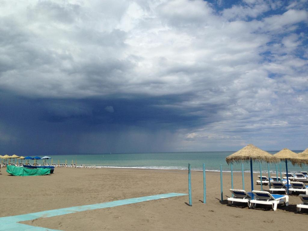 Pludmale ar smilšu krastu, ziliem sauļošanās krēsliem un mākoņainu debesu skatu virs ūdens.