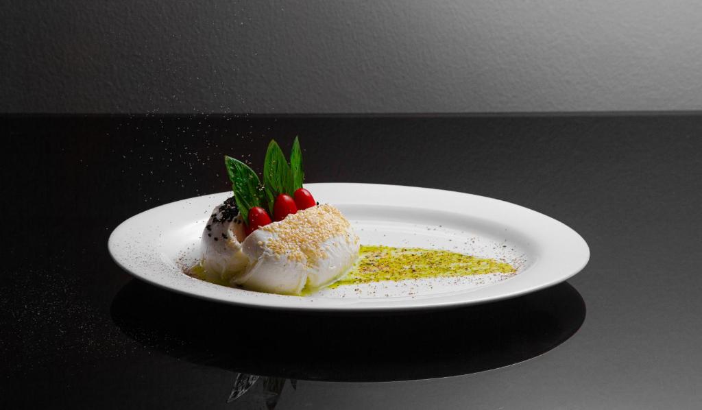 Gatavots ēdiens uz baltā šķīvja, dekorēts ar zaļām lapām un sarkaniem tomātiem, apbērts ar sezama sēklām un pistācijām.