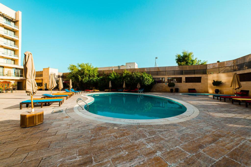 Peldbaseins ar sauļošanās krēsliem un lietussargiem, apkārt zaļiem augiem un viesnīcas ēku fons.