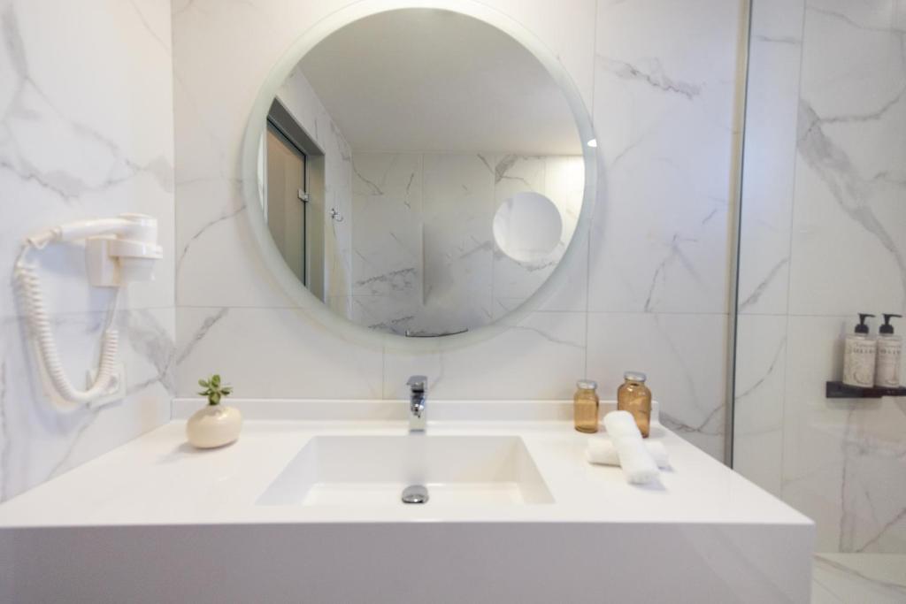 Mūsdienīgs vannas istabas interjers ar apaļu spoguli, baltu izlietni un dabīgu augu dekoru. Sienas ir klātas ar marmora raksta flīzēm, radot elegantu atmosfēru.