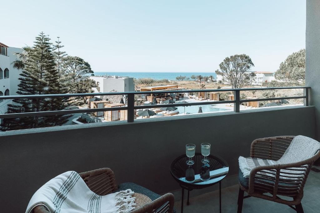 Balkons ar skatu uz jūru, divām ērtām krēsliem un galdiņu, uz kura atrodas dzērieni. Apkārt redzami zaļi koki un peldbaseins.