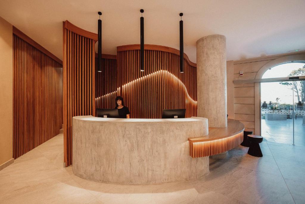 Mūsdienīga viesnīcas reģistratūra ar koka un betona elementiem, apgaismota ar stilīgām piekaramajām lampām. Receptionist, kas apkalpo viesus, radot aicinošu atmosfēru.