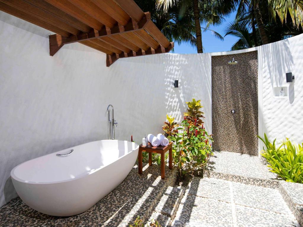 Atvērtā vannas istaba ar baltu brīvdabas vannu, akmens grīdu un tropiskajiem augiem, kas rada relaksējošu atmosfēru. Duša atrodas blakus, piedāvājot ērtu un modernu dizainu.