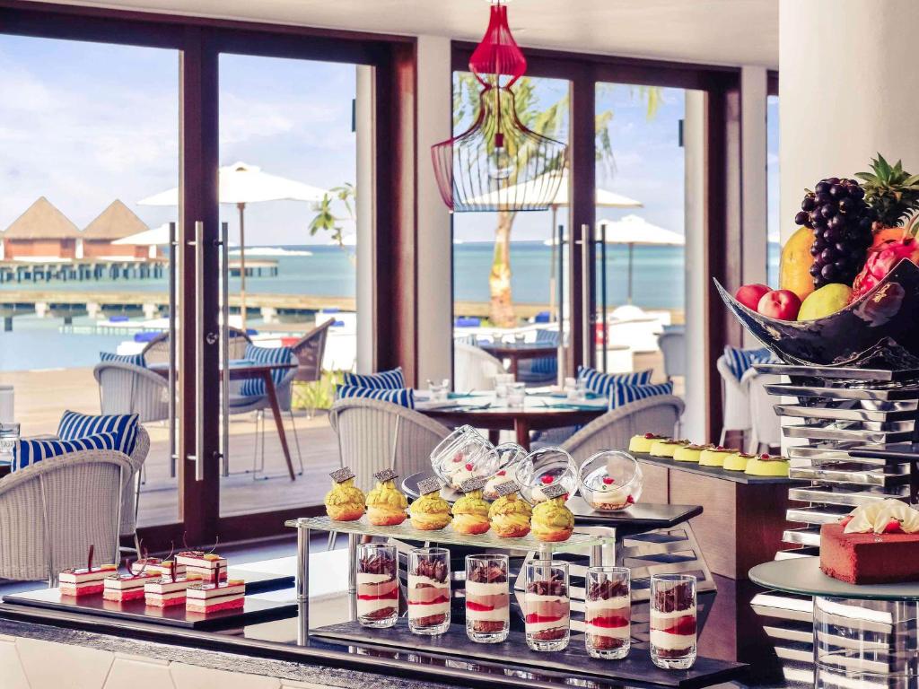 Elegants restorāns ar skatu uz jūru, kurā redzams izsmalcināts deserts un augļu grozs. Interjers ir gaumīgs ar komfortablām krēsliem un gaišiem toņiem, radot relaksējošu atmosfēru.