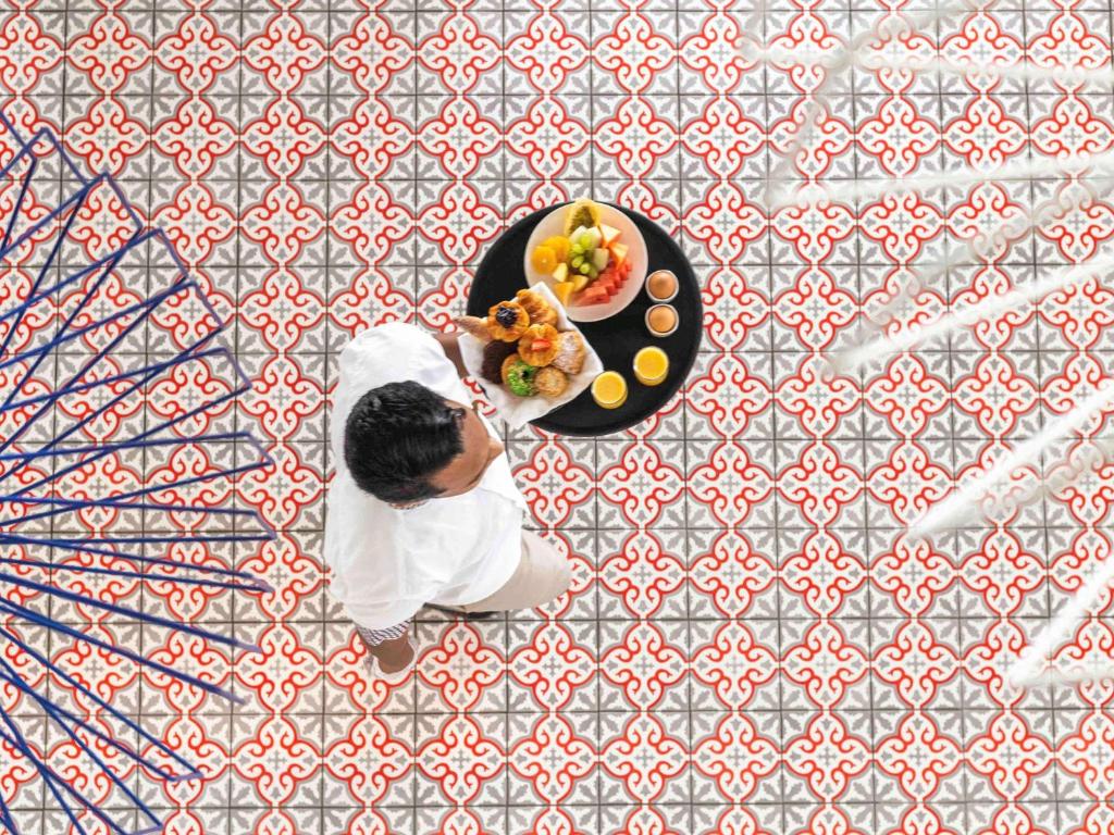 Vīrietis, kas nes pasniegšanas trauku ar augļiem un uzkodām, staigā pa greznu flīžu grīdu ar krāsainu rakstu.