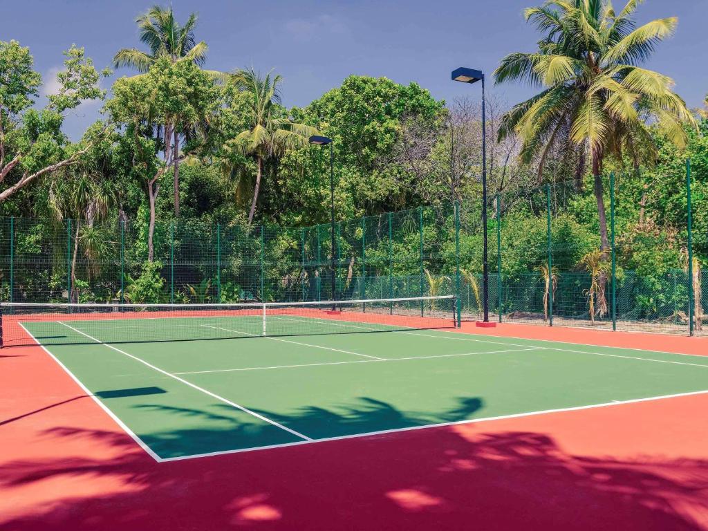 Tenisa laukums ar zaļu segumu un sarkanu maliņu, ieskauts tropiskā ainavā ar palmām un krūmiem. Ideāla vieta aktīvai atpūtai.