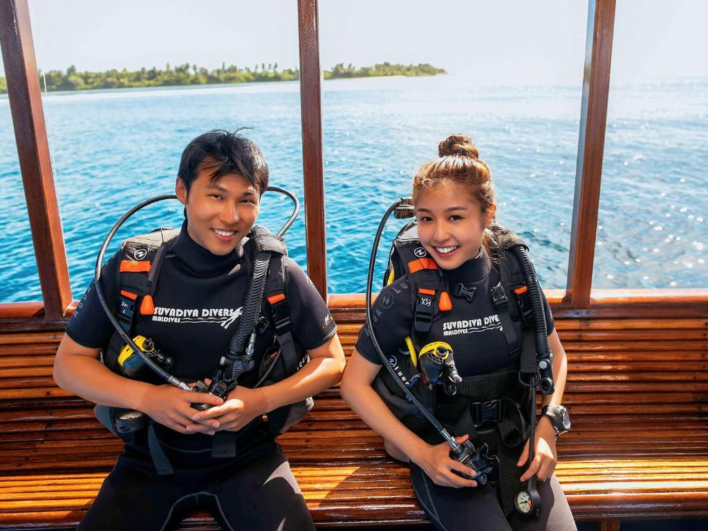 Divi jauni cilvēki, tērpušies niršanas ekipējumā, sēž uz laivas ar brīnišķīgu ūdens skatu fona. Viņi izskatās priecīgi un gatavi piedzīvojumiem zemūdens.