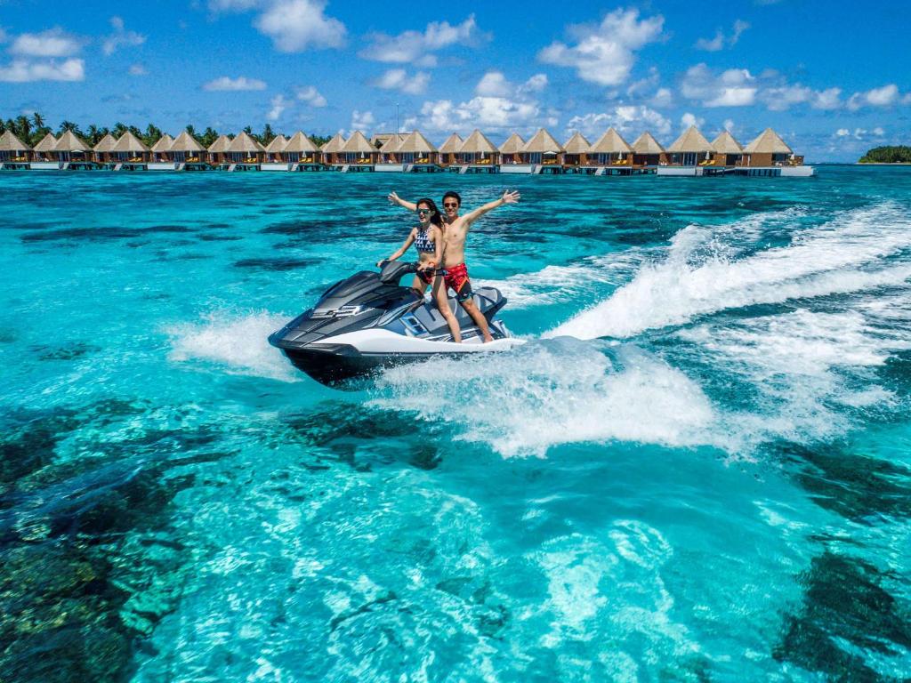 Jahtas brauciena prieks, divi cilvēki uz ūdens motocikla, fonā skaistas kūrorta mājas un dzidrs, zils ūdens.