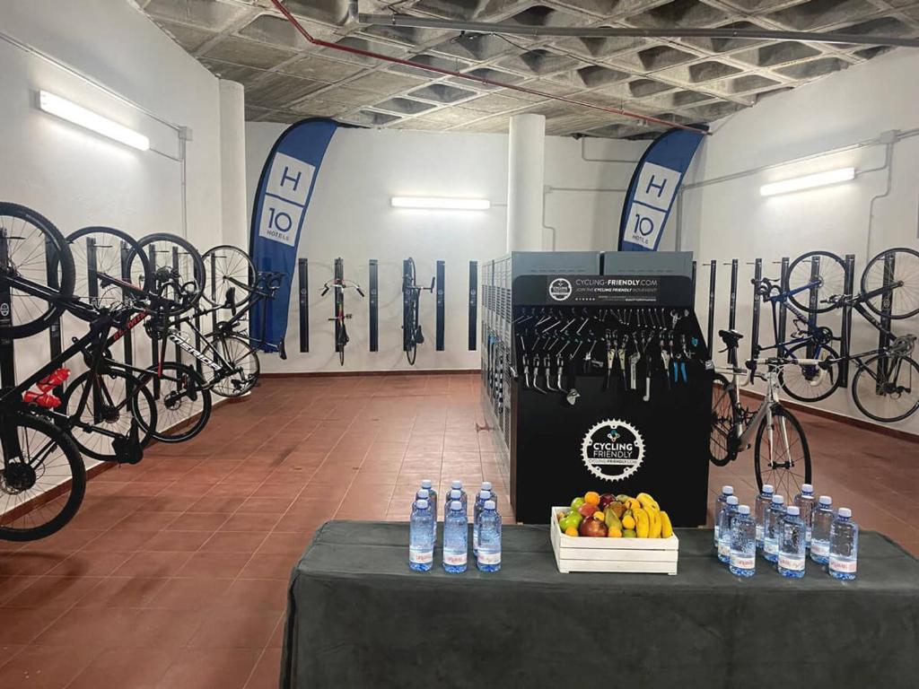 Velosipēdu uzglabāšanas telpa ar piekārtiem velosipēdiem, instrumentiem un uzkodu galdu, kas ietver augļus un ūdens pudeles. Telpa ir apgaismota un iekārtota, lai apkalpotu riteņbraucējus.