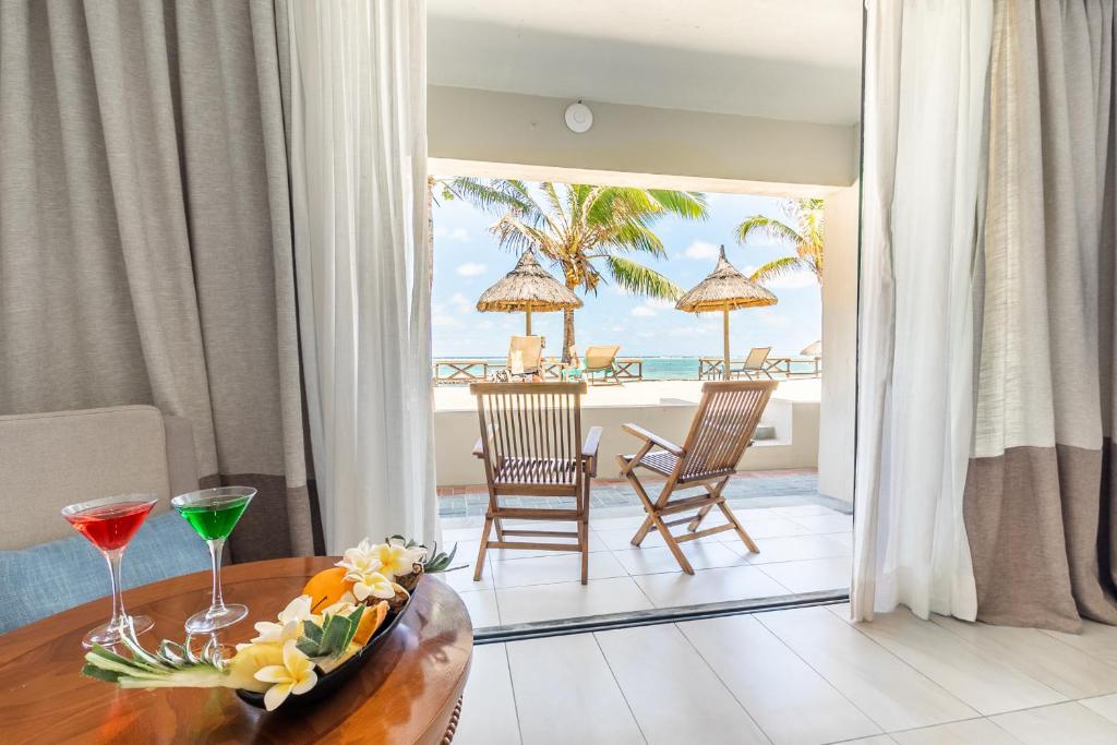 Izsmalcināta atpūtas telpa ar skatu uz pludmali, kurā redzami divi krēsli un palmas. Uz galda novietoti divi kokteiļi un ziedu kompozīcija.