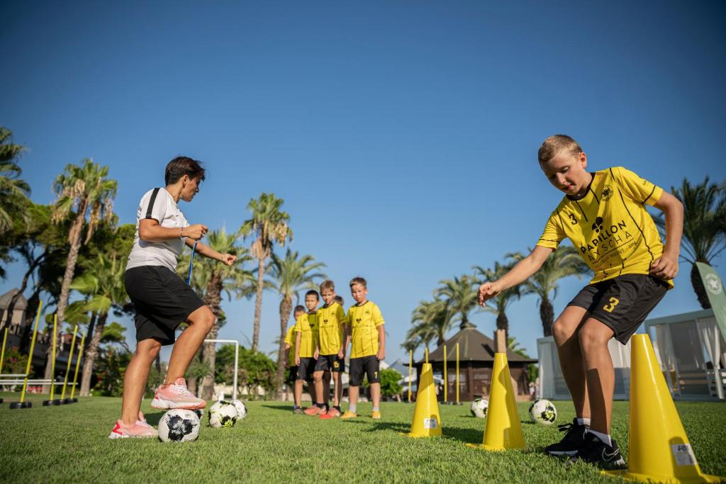 Bērni trenējas futbolā pie viesnīcas Papillon Ayscha, saulaina diena, zaļš zāliens.