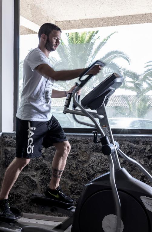 Vīrietis trenējas uz elipsveida trenažiera sporta zālē ar logu, caur kuru redz palmām apstādītu ainavu.