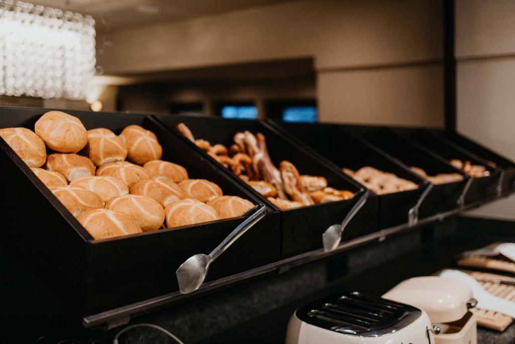 Maizes izstrādājumu bārs ar dažādām maizēm un konditorejas izstrādājumiem, kas sakārtoti melnās tvertnēs. Iekārtojums rada viesmīlīgu atmosfēru brokastu laikā.