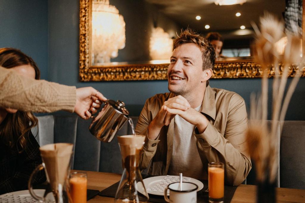 Vīrietis ar priecīgu izteiksmi sēž kafejnīcā, kamēr kāds lej kafiju no kannas. Blakus ir garšīgs dzēriens un kafijas pagatavošanas rīki. Interjers ir elegants ar zelta apdari un siltu apgaismojumu.