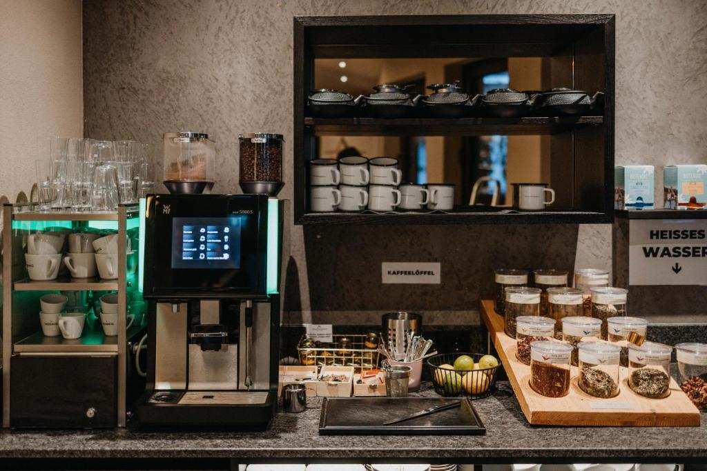 Kafijas stacija ar modernu kafijas automātu, glāzēm un dažādām tēju un garšvielu izvēlēm. Uz galda redzami arī augļi un piederumi dzērienu pagatavošanai.