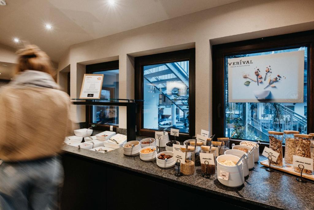 Brokažu galds ar dažādām graudaugu un riekstu izvēlēm, kas izvietots blakus logiem, caur kuriem redzams ārējais skats. Uz galda redzamas arī dažādas uzkodas un dekoratīvs plakāts.