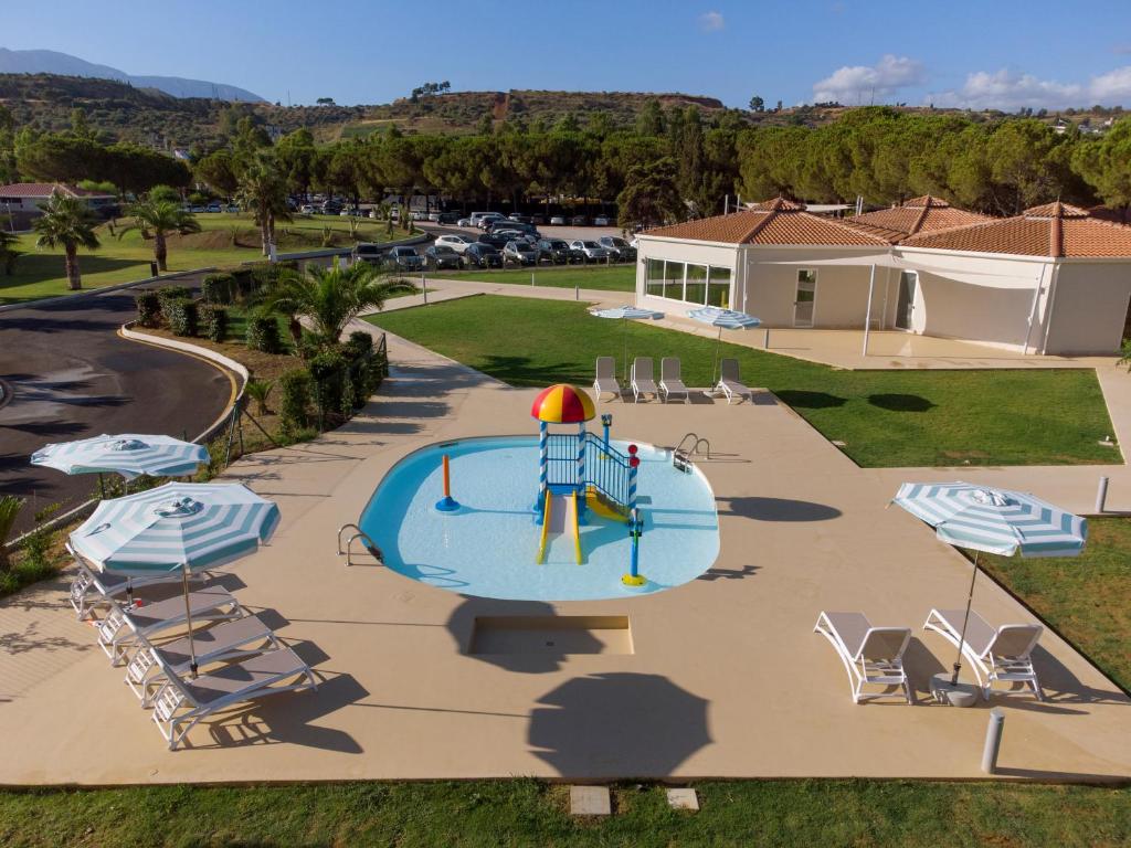 Bērnu baseins ar rotaļu struktūru un saulessargiem apkārt, kas atrodas zaļajā teritorijā. Apkārt ir koki un autostāvvieta, radot relaksējošu atmosfēru.