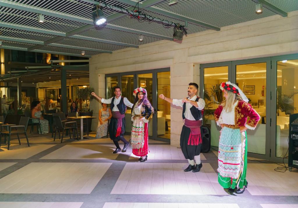 Dejotāji tradicionālās tērpos izpilda deju, kamēr apmeklētāji vēro pasākumu. Aiz dejotājiem redzama moderna restorāna vide ar ēdināšanas galdiem.