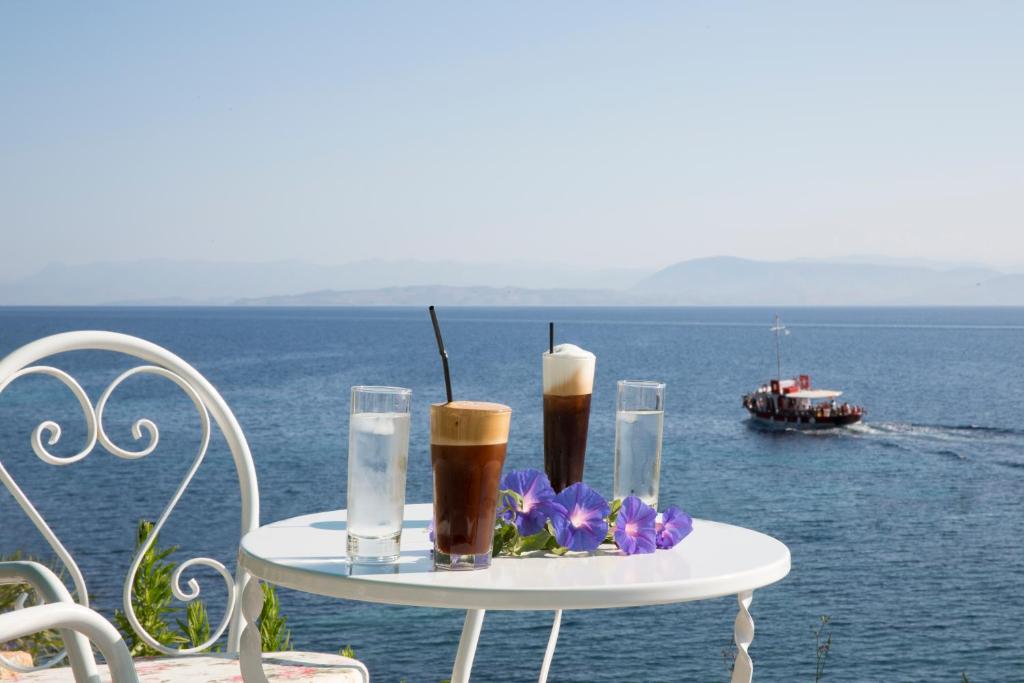 Vasaras dzērieni uz baltas galda piekrastes terasē ar skatu uz jūru un laivu. Ziedi pie galda piešķir noskaņu atpūtai.