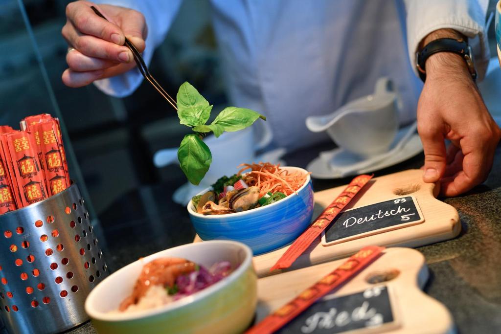 Šefpavārs izmanto dakšiņu, lai pievienotu baziliku pie ēdiena, kam blakus ir divas bļodas ar nūdeles un garnelēm. Uz galda ir redzami arī ēdiena zīmes un pāris kociņi.