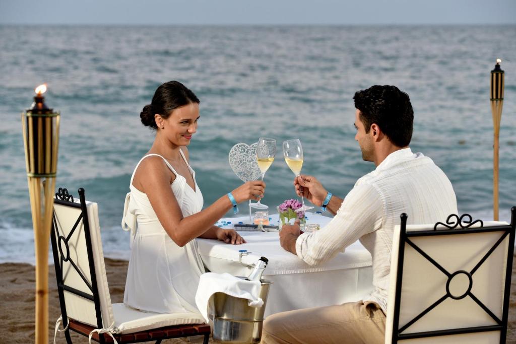Romantiska vakara maltīte pie jūras ar diviem cilvēkiem, kas cīnās ar šampanieti, sēžot pie elegantas galda ar pludmales ainavu fonā.