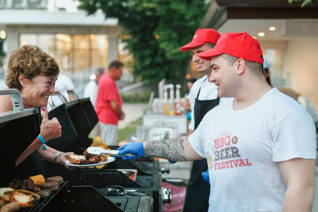 Siltā atmosfērā notiek BBQ festivāls, kur cilvēki izbauda grilētu ēdienu. Sieviete ar laimīgu izteiksmi saņem ēdienu no grilētāja, kurš valkā T-kreklu ar festivāla uzrakstu.