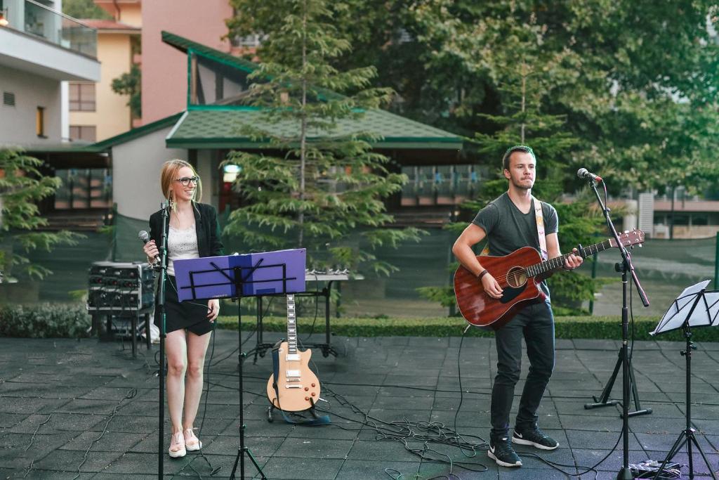 Mūziķi uzstājas ārā, viens ar akustisko ģitāru, otrs ar mikrofonu, sagatavojoties koncertam. Apkārt ir zaļš fons un mūzikas aprīkojums.