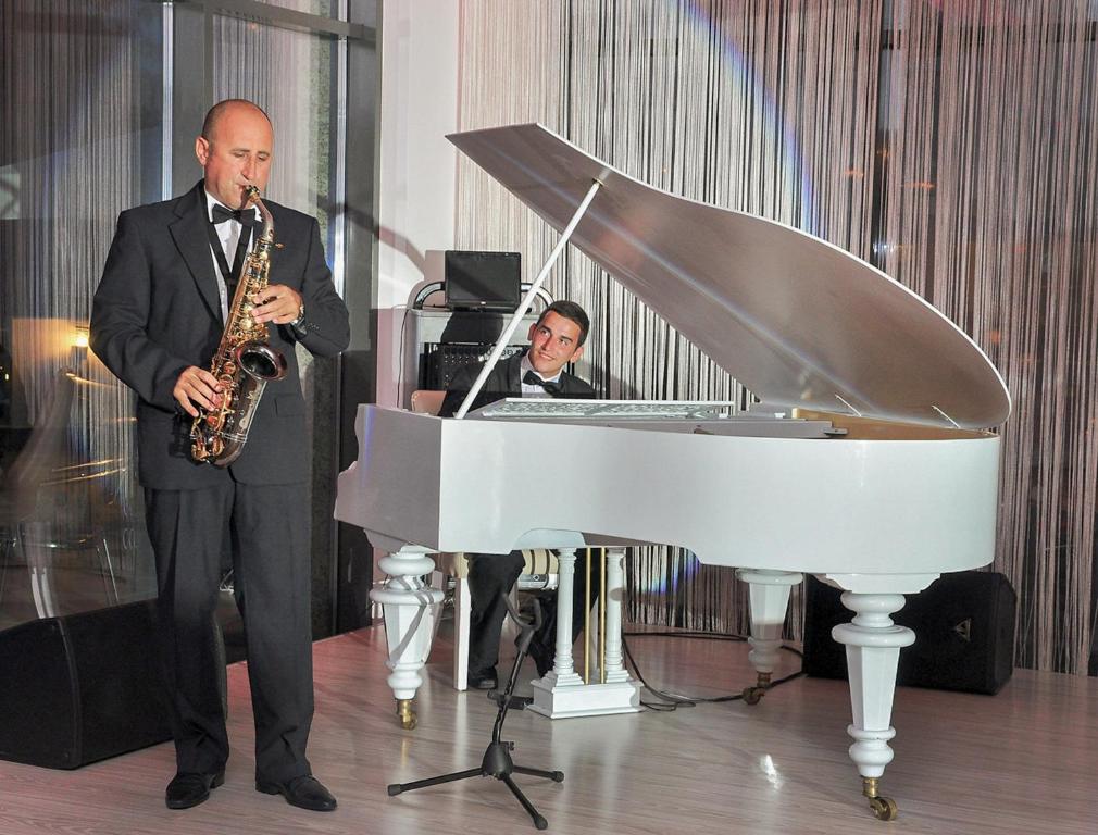 Divi mūziķi izpilda dzīvās mūzikas priekšnesumu, viens spēlē saksofonu, bet otrs sēž pie balta akustiskā klavieru instrumenta.