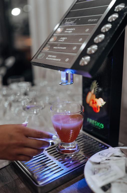 Kokteiļu automāts ar digitālo paneli, no kura tiek pildīts dzēriens glāzē. Fonā redzami tukši glāzes un šķīvītis ar saldumiem.