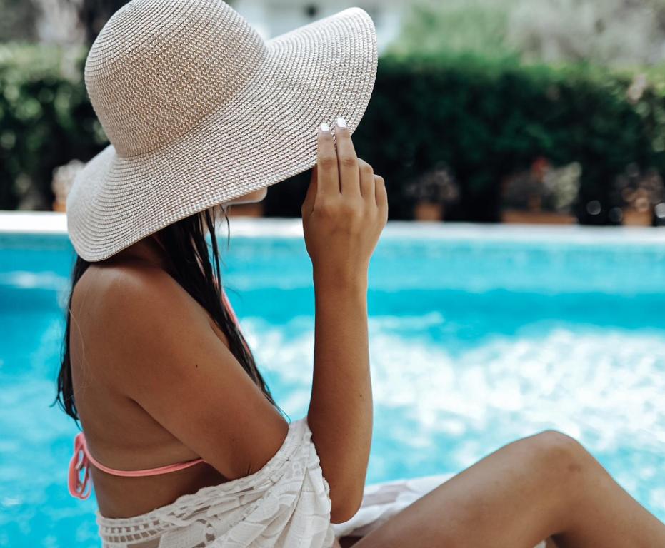 Dāma ar lielu saules cepuri sēž pie peldbaseina, turot roku pie cepures. Apkārt ir zaļa vide un dzidrs ūdens.