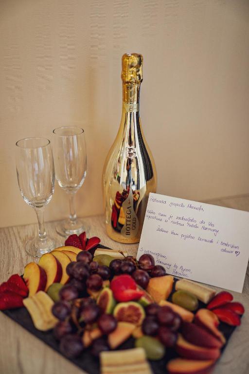 Zeltaina šampanieša pudele ar divām glāzēm un augļu šķīvis, kas ietver dažādas svaigas ogas un augļus, papildināts ar apsveikuma kartiņu.