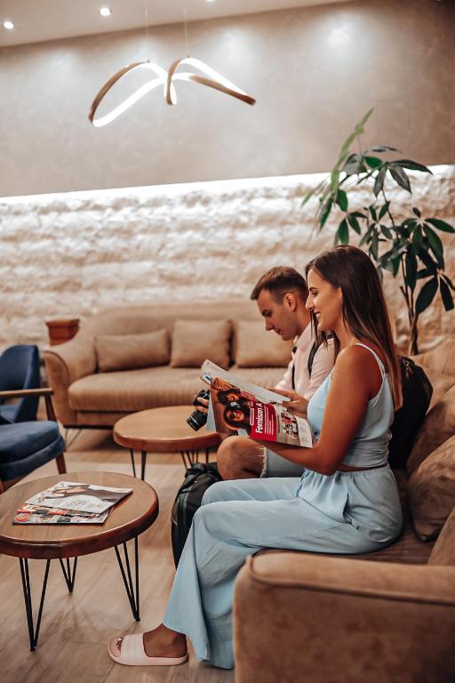 Jaunā sieviete sēž uz dīvāna un lasa žurnālu, kamēr vīrietis blakus tur kameru. Interjers ir moderns ar dabīgām krāsām un augiem, radot relaksējošu atmosfēru.