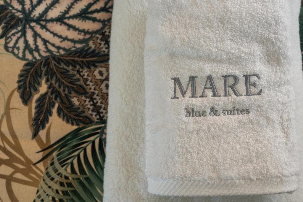 Balts dvielis ar izšūtu nosaukumu "MARE blue & suites" uz fona ar zaļām lapām un ziediem.