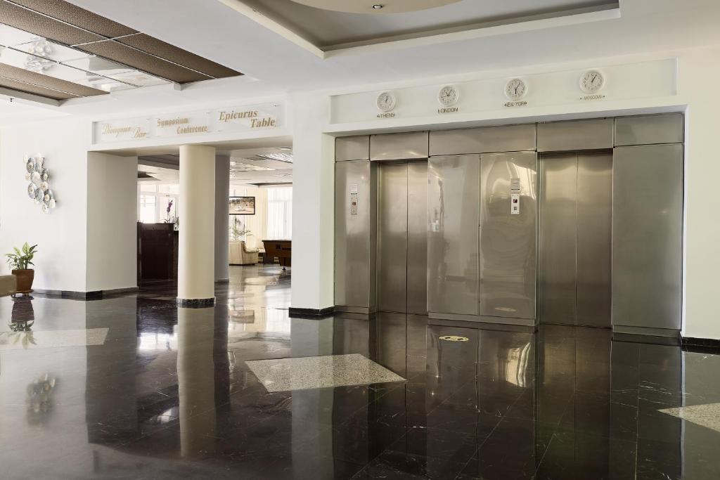 Mūsdienīgs viesnīcas vestibils ar spīdīgām melnām grīdām, kurā redzami divi lifti un ieeja uz konferenču telpām. Telpas interjers ir gaišs un elegants, radot viesmīlīgu atmosfēru.