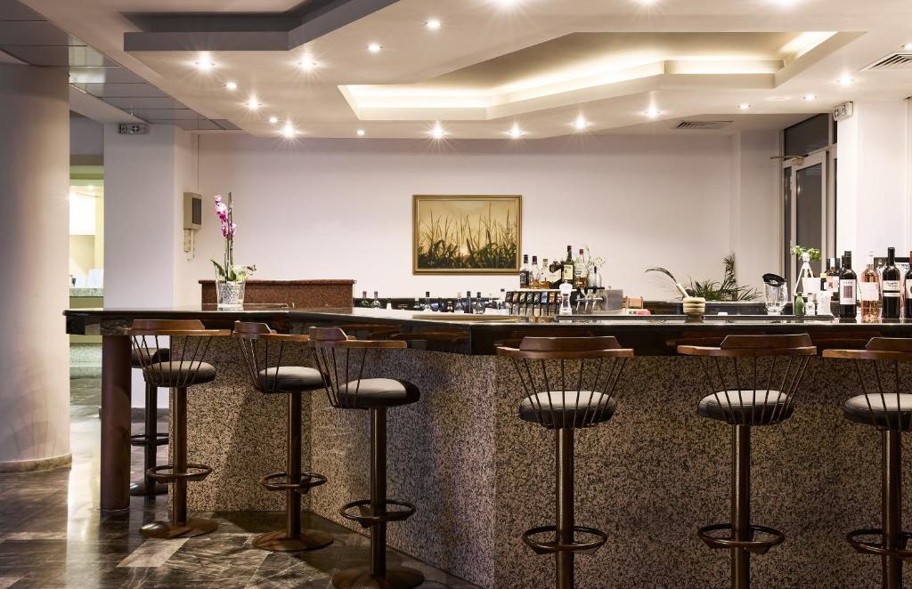 Mūsdienīgs bārs ar elegantu akmens virsmu, koka krēsliem un izsmalcinātu apgaismojumu, kas rada patīkamu atmosfēru. Uz bāra plaukta ir izstādes ar dažādiem dzērieniem un dekoratīvām augu kompozīcijām.