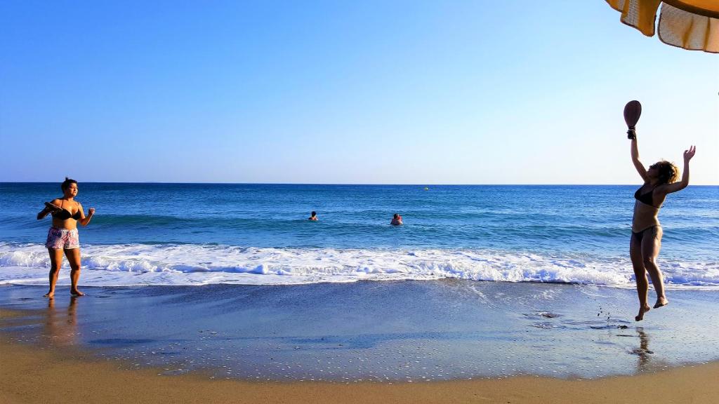 Divas sievietes spēlē pludmales spēli pie jūras, kamēr citi peld ūdenī. Skaists saulains laiks un skaidrs debesis.