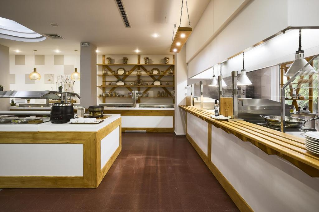 Mūsdienīgs restorāna interjers ar koka un baltām detaļām, izceļot plašu un gaišu telpu. Skats uz bufetes staciju un atvērtu virtuvi ar stilīgu apgaismojumu.