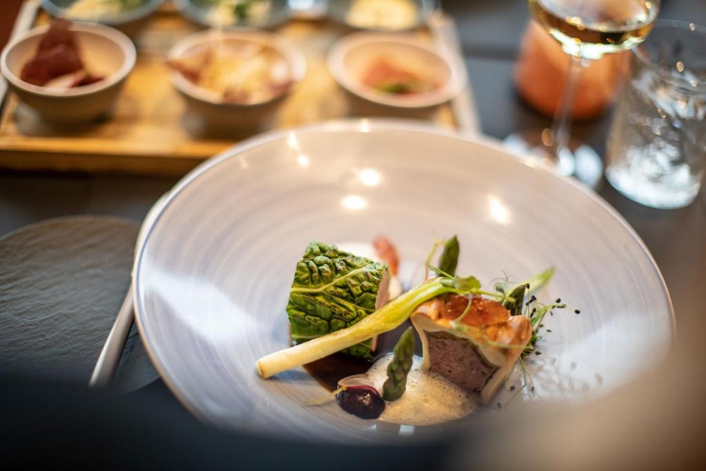 Eleganta plāksne ar izsmalcinātu ēdienu, kas satur gaļas gabalu ar dārzeņu garnējumu un mērci. Fonā redzami mazi trauki ar dažādiem uzkodām un vīns.