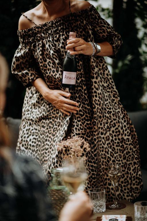 Sieviete leopardu raksta kleitā, turētā pudelē šampanieša "Ayala", svinīgā atmosfērā. Galda priekšplānā redzami dzērieni un dekorācijas.