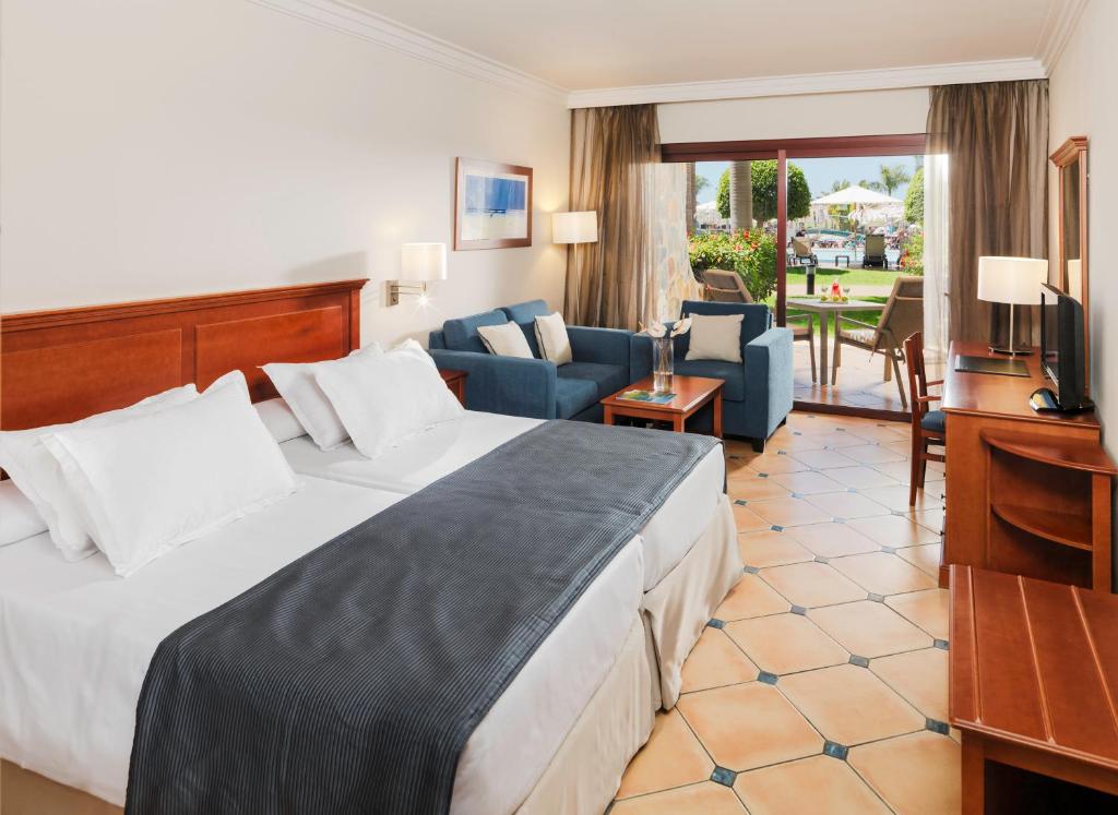 Mūsdienīga viesnīcas numura iekārtojums ar divguļamo gultu, dīvānu un skatu uz dārzu. Numurā ir arī rakstāmgalds un televizors, radot komfortablu atmosfēru atpūtai.