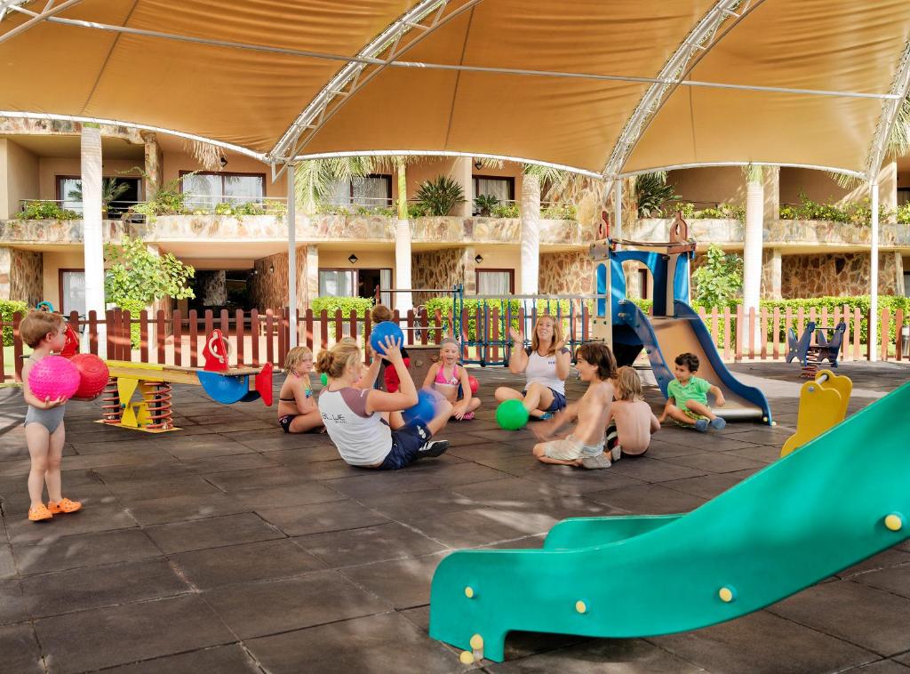 Bērni spēlējas rotaļu laukumā zem jumta, izmantojot krāsainas bumbas un izklaidējoties pie slide. Apkārt ir dažādi rotaļu elementi un zaļumi, radot draudzīgu un jautru atmosfēru.