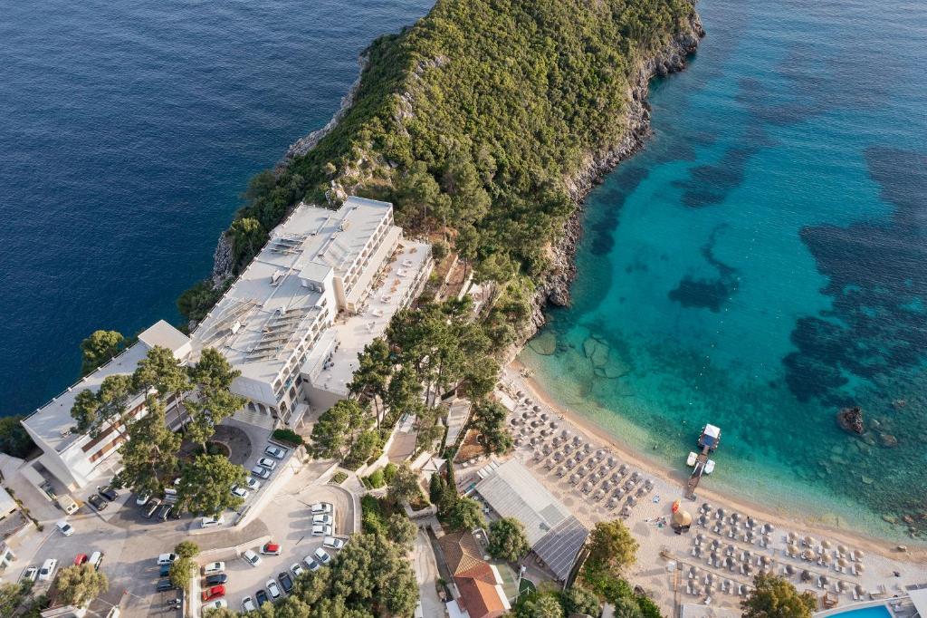 Viedoklis no gaisa uz viesnīcu pie jūras, kur blakus ir pludmale ar sauļošanās krēsliem un skaidra ūdens. Apkārtne ir apzaļumota ar kokiem un klintīm, radot mierīgu atmosfēru.