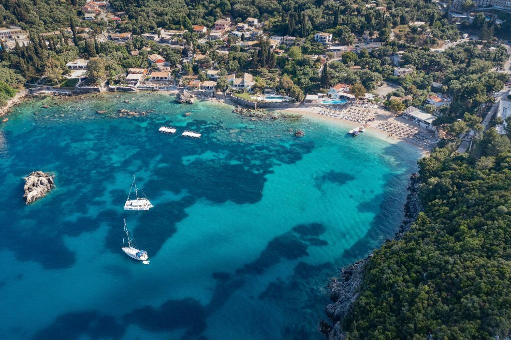 Kopts skats uz krāšņu pludmali un dzidru zilo ūdeni, kur kuģi peldoši atrodas netālu no krasta. Apkārt redzami zaļi koki un māju jumti, radot mierīgu un idillisku ainavu.