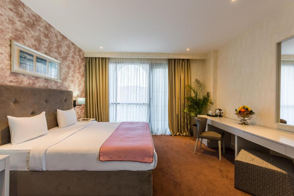 Mūsdienīga viesnīcas numura iekārtojums ar plašu gultu, elegantiem aizkariem un viesmīlīgu atmosfēru. Interjerā ir redzama arī dekoratīva auga un augļu kompozīcija.