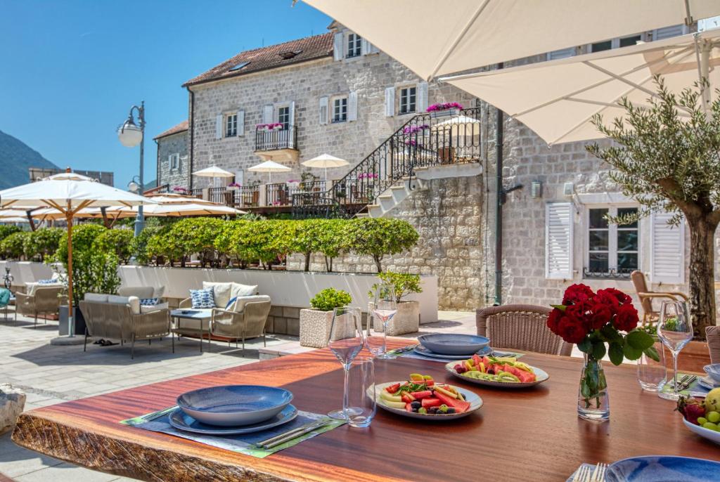 Elegants restorāna terase ar malkošanas galdu, uz kura ir krāsaini augļi un ziedi, apkārtējo ainavu papildina vēsturiskas ēkas un saulainas parasoles.