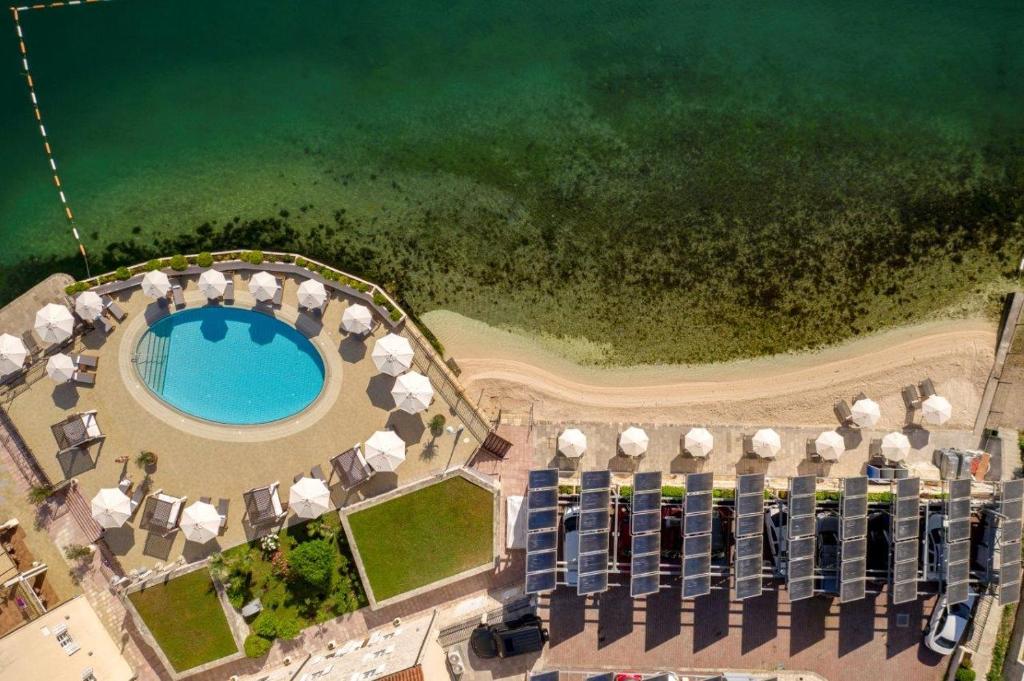 Peldbaseins ar saulessargiem un pludmali, redzams no augšas, ar zaļganām ūdenstilpnēm un saules paneļiem.