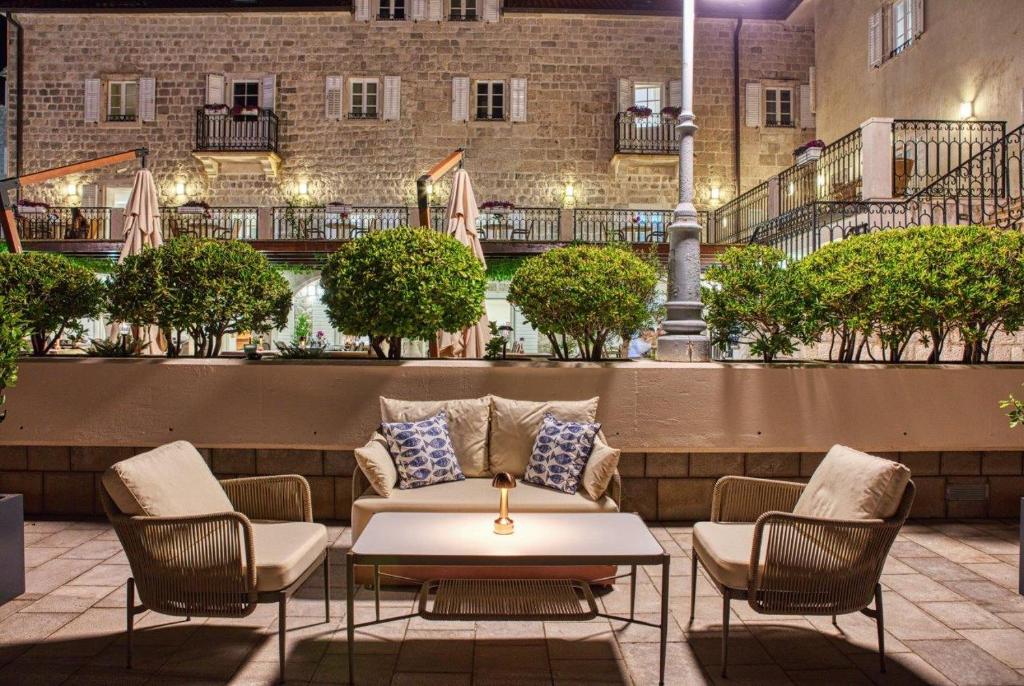 Mūsdienīgs āra atpūtas stūris ar ērtām krēslām un galdu, apzaļumots ar podiņiem un apgaismots naktī. Fons veido vēsturiska akmens ēka ar balkoniem un terasēm.