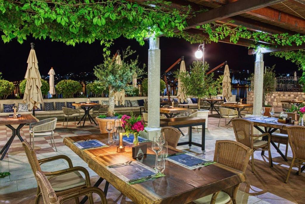 Eleganta āra restorāna terase ar koka galdiem, ziediem un apgaismojumu, kas rada patīkamu atmosfēru. Nakts ainava ar zaļumu un ēdamzālēm pie ūdens.
