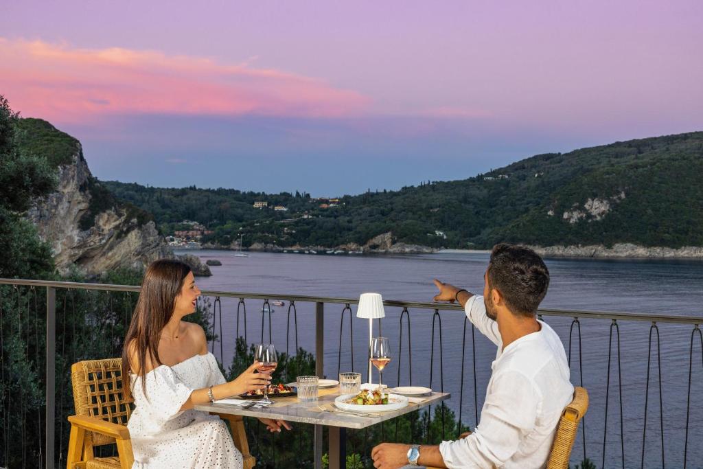 Romantiska vakariņas ar skatu uz jūru, kur pāris izbauda maltīti un vīnu, kamēr vēro skaisti saulrieta toņus.