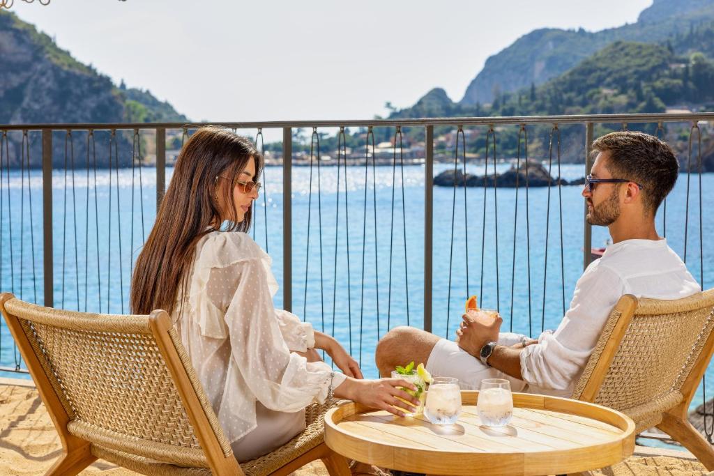 Jauns pāris atpūšas pie jūras, baudot kokteiļus saulainā dienā. Viņi sēž uz krēsliem, baudot skatu uz ūdeni un apkārtējo dabu.